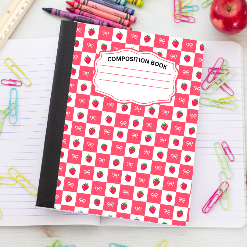 A composition book with a red and white checkered cover pattern featuring small strawberry and bow illustrations, placed on top of an open lined notebook with colorful paperclips, crayons, and an apple around it.