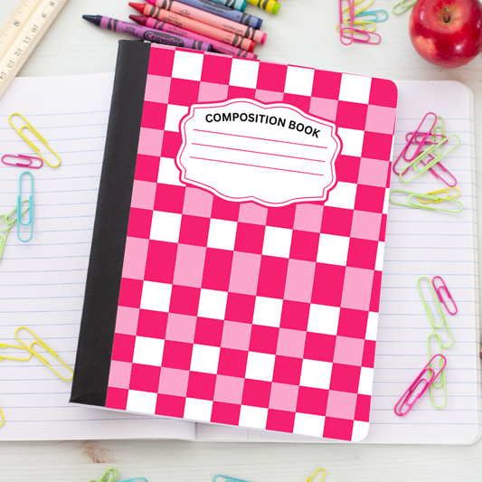 A pink and white checkered composition book with a black spine, placed on an open lined notebook surrounded by school supplies like crayons, paper clips, a ruler, and an apple.