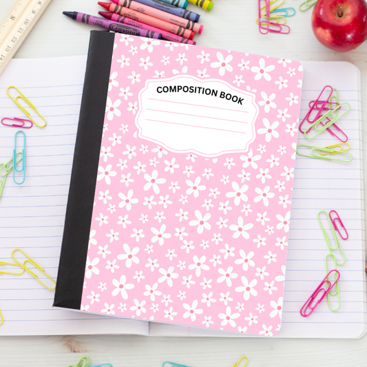 A pink composition book with a white floral pattern and a black spine, placed on an open lined notebook surrounded by colorful paperclips, crayons, a ruler, and an apple.
