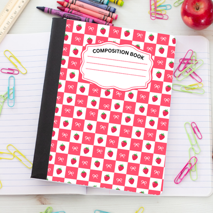 A composition book with a red and white checkered cover pattern featuring small strawberry and bow illustrations, placed on top of an open lined notebook with colorful paperclips, crayons, and an apple around it.