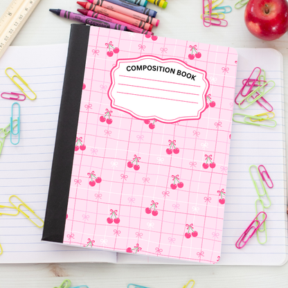 A pink composition notebook with a cherry pattern and a black spine, placed on an open lined notebook surrounded by colorful paperclips, crayons, a ruler, and an apple.