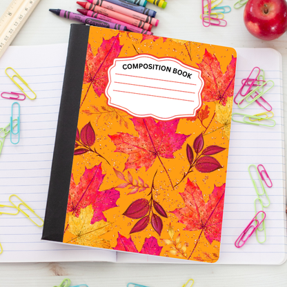 A composition notebook with a bright orange autumn leaf cover pattern, placed on an open lined notebook surrounded by colorful paperclips, crayons, a ruler, and an apple.