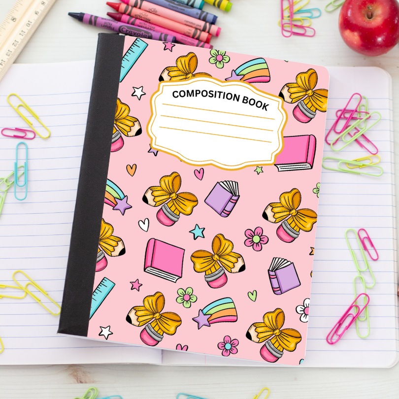 A pink composition book with a black spine, decorated with colorful school-themed illustrations including yellow bows, pencils, stars, flowers, and books. The book is placed on an open lined notebook surrounded by paperclips, crayons, a ruler, and an apple.