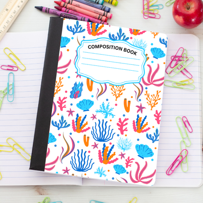 A composition book with a colorful underwater coral pattern on a white cover, placed on an open lined notebook surrounded by paperclips, crayons, and an apple.