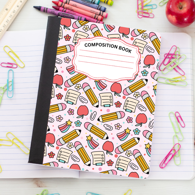 A composition notebook with a pink cover decorated with cartoon pencils, apples, stars, and writing tools, placed on top of an open lined notebook with colorful paperclips around it.