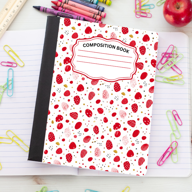 A composition notebook with a white cover decorated with a red strawberry and berry pattern, placed on an open lined notebook surrounded by colorful paperclips, crayons, and an apple.