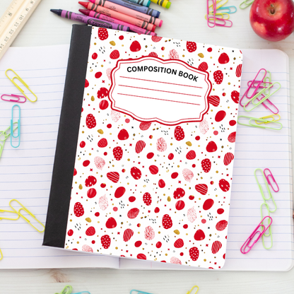 A composition notebook with a white cover decorated with a red strawberry and berry pattern, placed on an open lined notebook surrounded by colorful paperclips, crayons, and an apple.