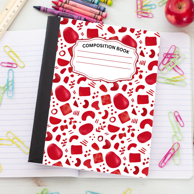 A composition book with a white cover decorated with a red pattern of apples and geometric shapes, placed on an open lined notebook surrounded by colorful paperclips, crayons, and an apple.