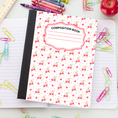 A composition book with a white cover decorated with a red and green cherry pattern, featuring a black spine and a label that reads 'COMPOSITION BOOK'. It is styled on a white desk with school supplies including colored paperclips, crayons, a ruler, and an apple.