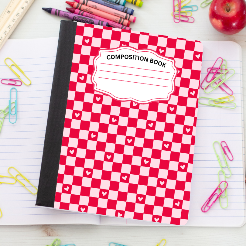 A red and white checkered composition book with a black spine, placed on an open lined notebook surrounded by colorful paperclips, crayons, a ruler, and an apple.