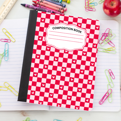 A red and white checkered composition book with a black spine, placed on an open lined notebook surrounded by colorful paperclips, crayons, a ruler, and an apple.