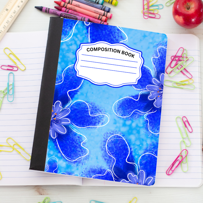 A blue floral composition notebook with a black spine, placed on an open lined notebook surrounded by colorful paperclips, crayons, a ruler, and an apple.
