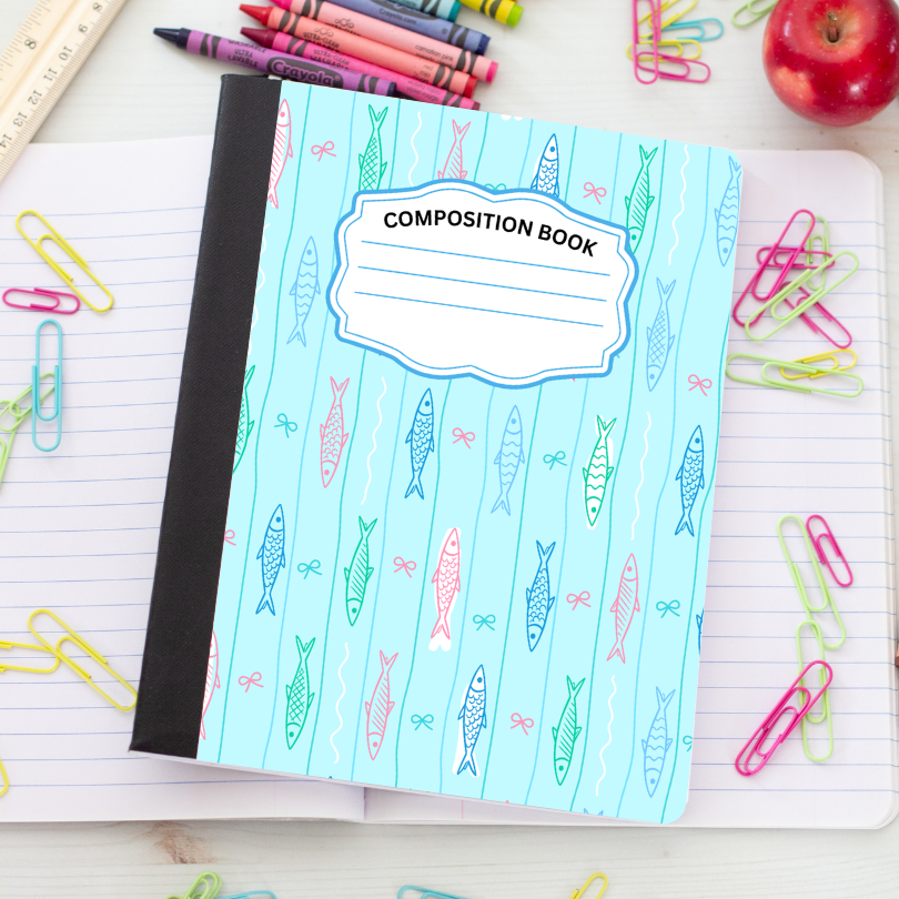 A composition book with a light blue cover patterned with colorful fish and bows, placed on an open lined notebook surrounded by school supplies like paper clips, crayons, a ruler, and an apple.