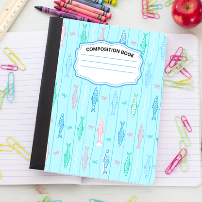 A composition book with a light blue cover patterned with colorful fish and bows, placed on an open lined notebook surrounded by school supplies like paper clips, crayons, a ruler, and an apple.