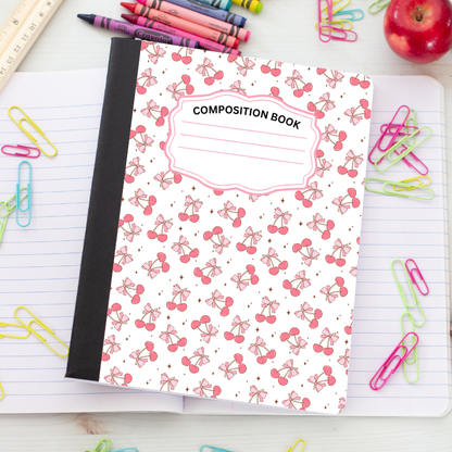 A composition notebook with a white cover decorated with a pink cherry and bow pattern, a black spine, and a label reading 'COMPOSITION BOOK'. It is styled on an open lined notebook surrounded by school supplies like paper clips, crayons, and an apple.