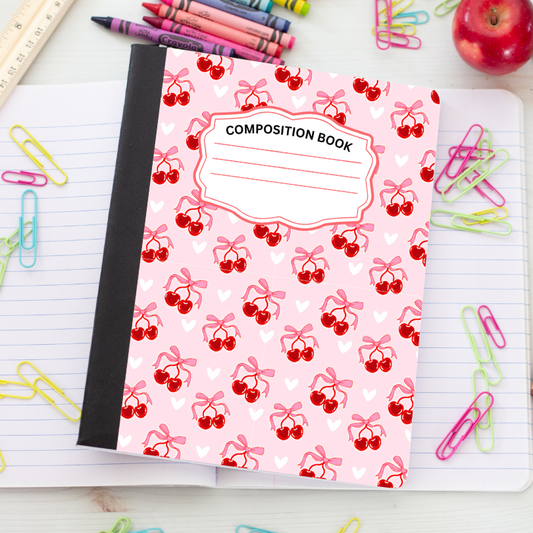 A pink composition notebook with a cherry and heart pattern, placed on a lined desk with school supplies like crayons, paper clips, and an apple.