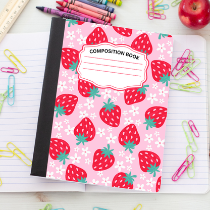 A pink composition notebook with a strawberry and white flower pattern, featuring a black spine and a blank label on the cover, styled on a white desk with stationery items.