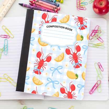 A composition book with a light blue and white gingham cover pattern featuring red lobsters, yellow lemon slices, and blue bows, displayed on a lined notebook surrounded by school supplies.